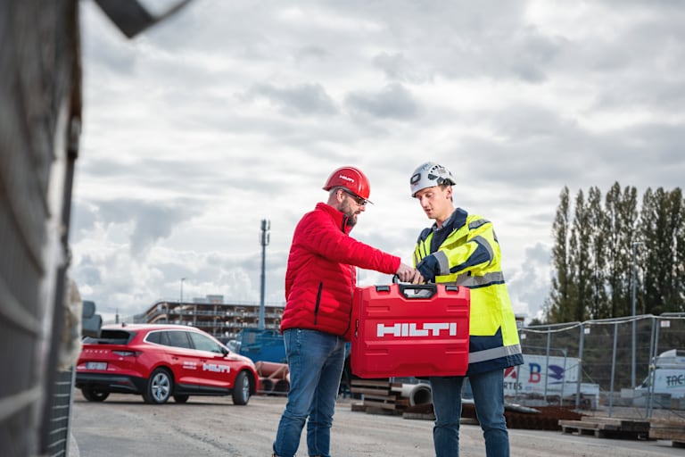 Un commercial Hilti échange avec un client Hilti sur un chantier. Un véhicule électrique Hilti est visible en arrière-plan.