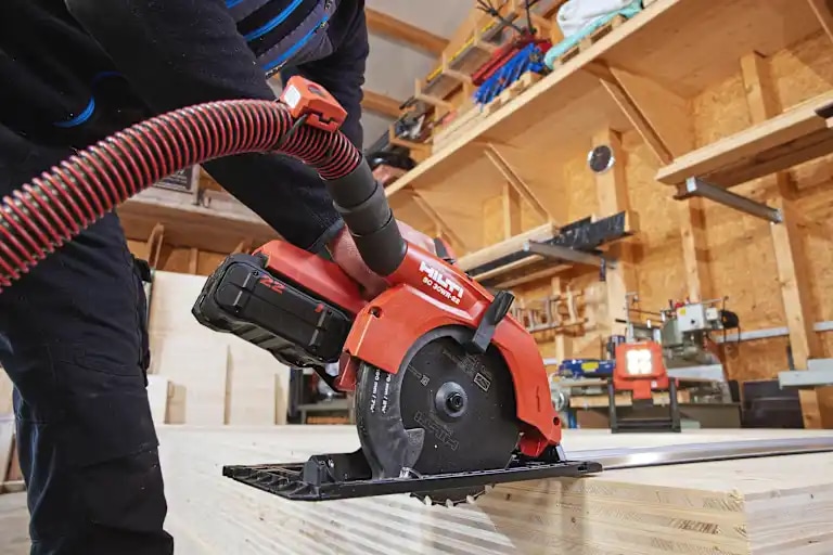 Worker is cutting a CLT plate with a circular saw SC 30WR-22, a cordless optimized wood blade and a guide rail. In the background the jobsite lamp SL 6-22 illuminates the scenerie. The circular saw is connected to a vaccuum cleaner
