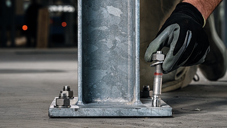 Hand mit Arbeitshandschuh setzt einen Hilti Bolzenanker HST4 in die Bodenplatte eines Stahlträgers ein, um ihn im Betonboden zu verankern