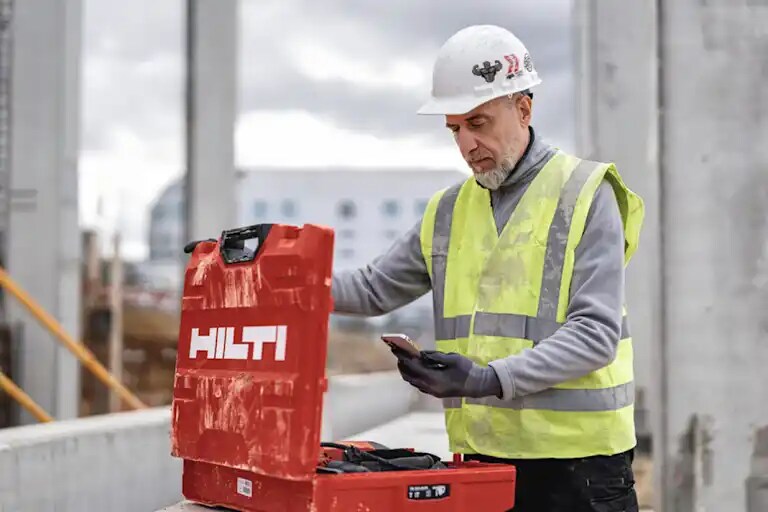 Un trabajador en una obra sostiene un teléfono en la mano, junto a una caja Hilti abierta.