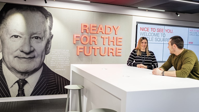 Dois membros da equipa Hilti reunidos num espaço comum, com o mural da cultura e valores Hilti no escritório de Manchester.
