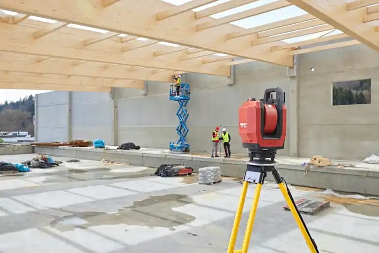A total station PLT 400 in a construction site hall with wooden roof. In the background some jobsite workers and a Hilti representative