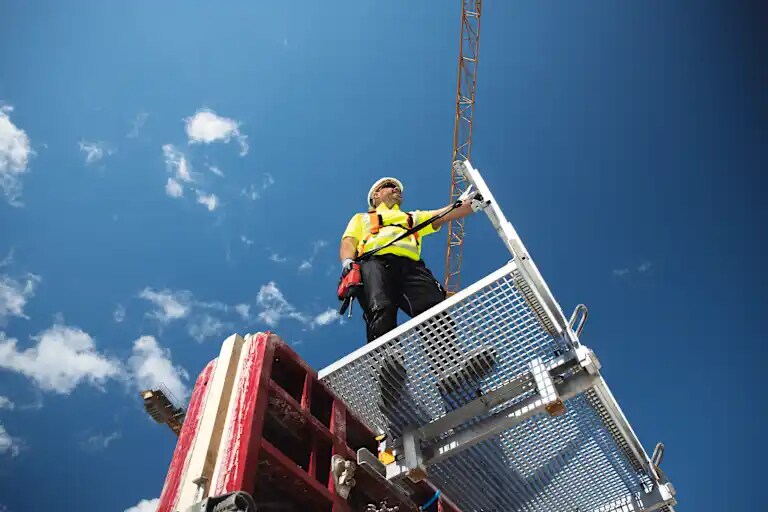Jobsite manager with tether and TE 6-22 cordless rotary hammer standing on scaffold at height overlooking construction site. Cranes and blue sky in the background.