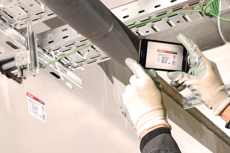Zwei Hände in Schutzhandschuhen, die ein Handy halten, auf dem Display ist ein Prüfaufkleber zu sehen, im Hintergrund die Schienen und Rohre einer Baustelle.