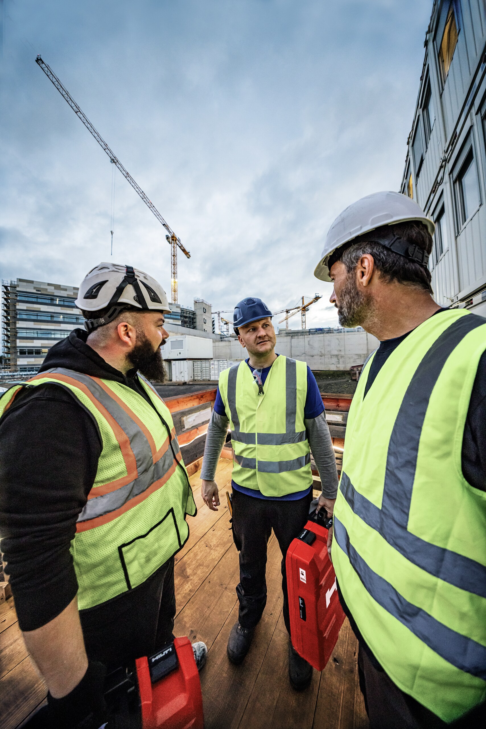 Three male workers in conversation on jobsite, carrying Hilti cases