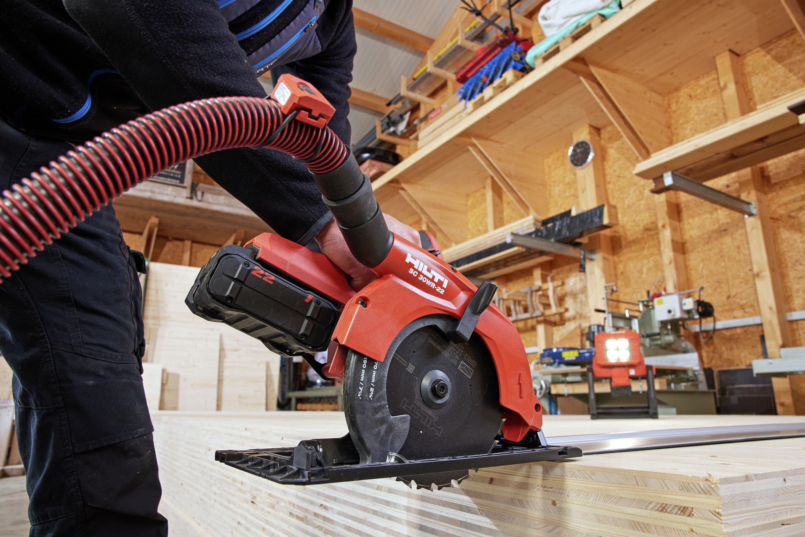 Worker is cutting a CLT plate with a circular saw SC 30WR-22, a cordless optimized wood blade and a guide rail. In the background the jobsite lamp SL 6-22 illuminates the scenerie. The circular saw is connected to a vaccuum cleaner