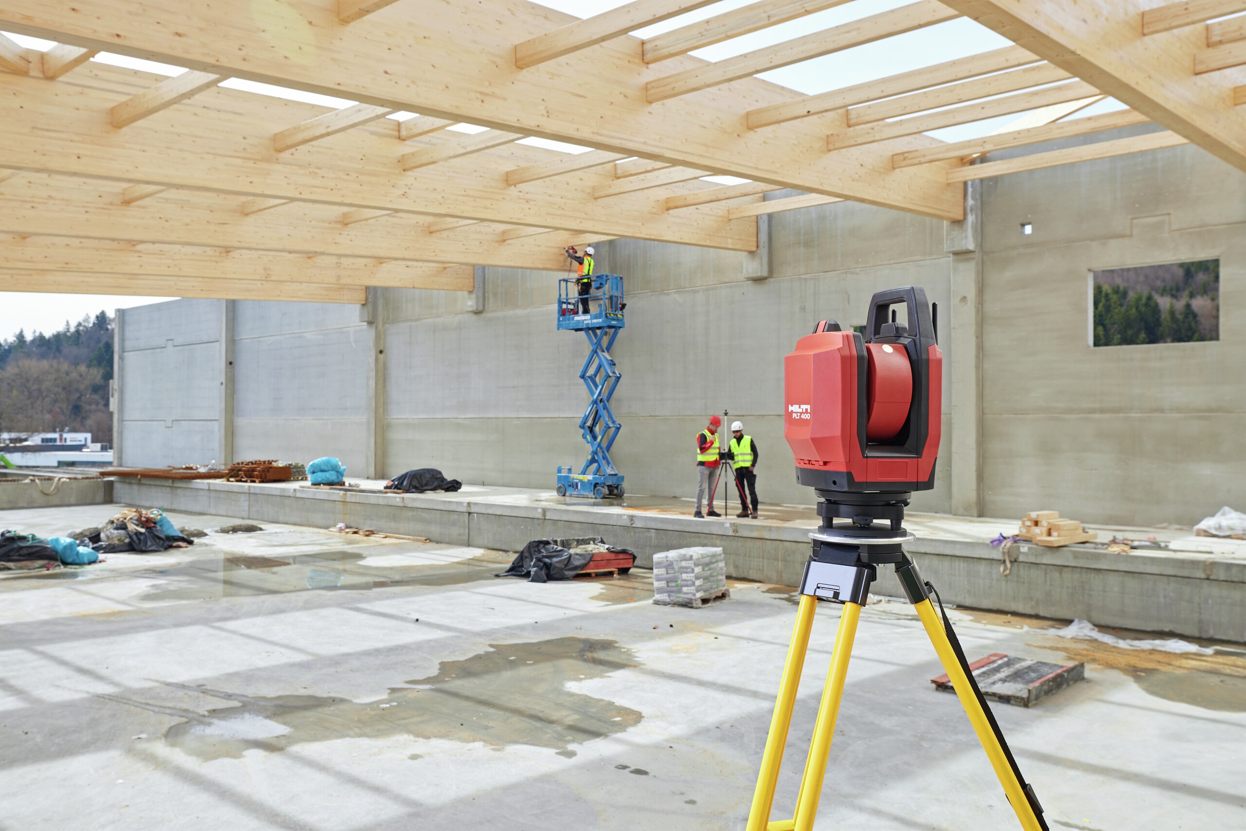 A total station PLT 400 in a construction site hall with wooden roof. In the background some jobsite workers and a Hilti representative