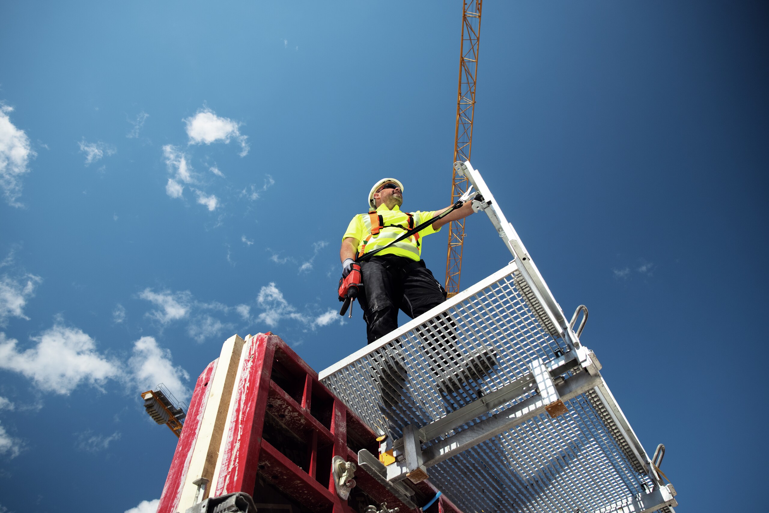 Jobsite manager with tether and TE 6-22 cordless rotary hammer standing on scaffold at height overlooking construction site. Cranes and blue sky in the background.
