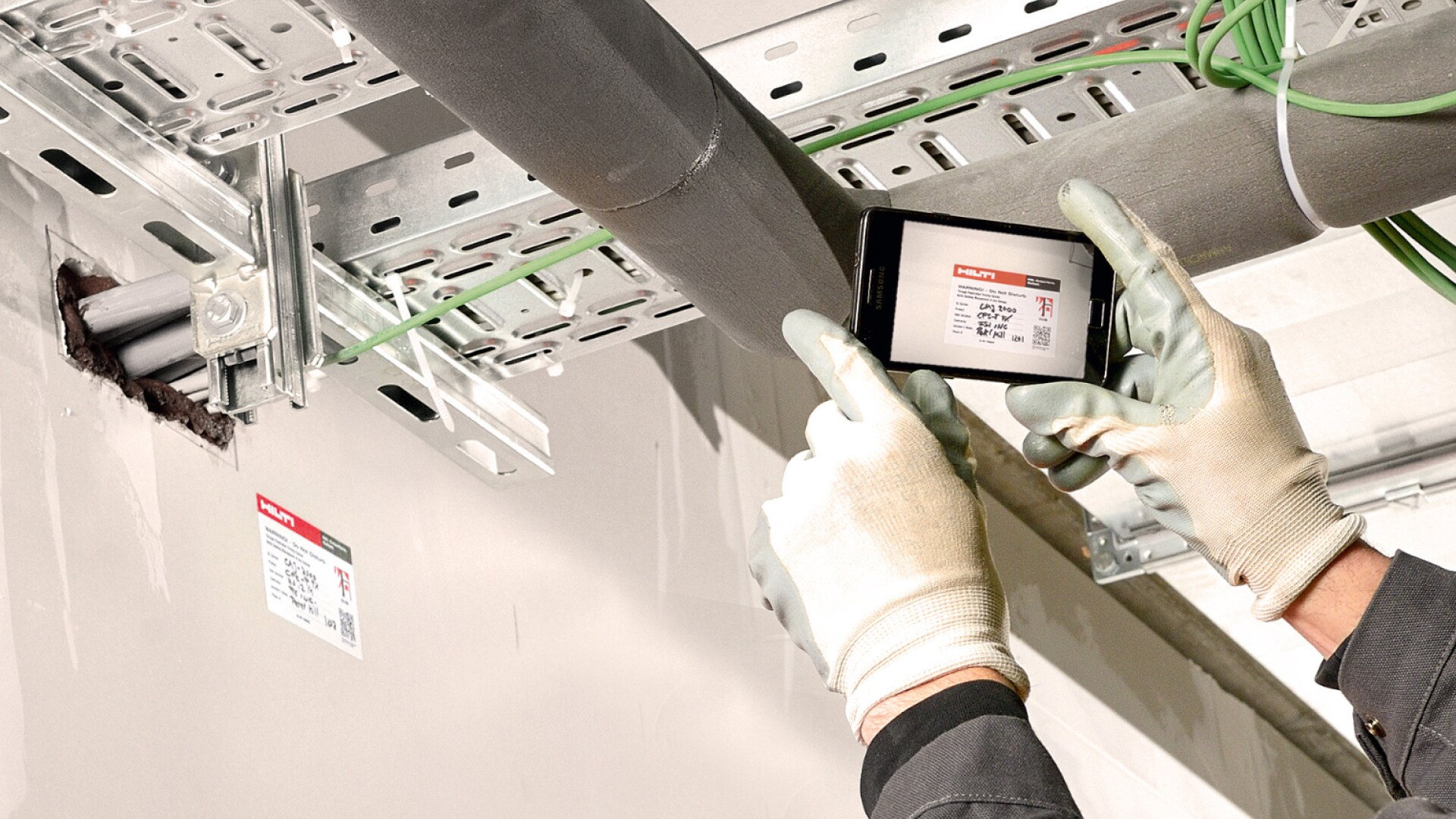 Zwei Hände in Schutzhandschuhen, die ein Handy halten, auf dem Display ist ein Prüfaufkleber zu sehen, im Hintergrund die Schienen und Rohre einer Baustelle.